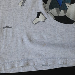 May include: Gray t-shirt with a graphic of a soccer ball with blue and white accents. The text "NUTMEG" is printed on the bottom left corner of the shirt.