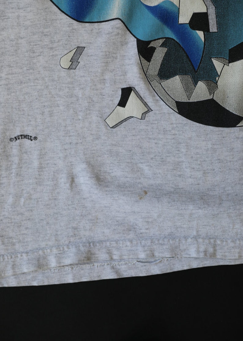 May include: Gray t-shirt with a graphic of a soccer ball with blue and white accents. The text "NUTMEG" is printed on the bottom left corner of the shirt.