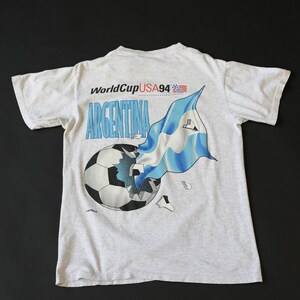 May include: A gray t-shirt with a graphic of a soccer ball being broken by an Argentinian flag. The text "World Cup USA 94" and "Argentina" are printed on the shirt.