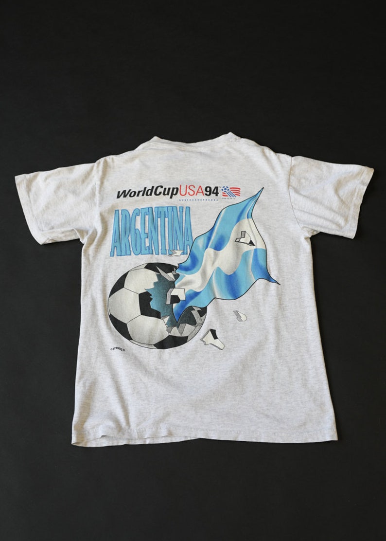 May include: A gray t-shirt with a graphic of a soccer ball being broken by an Argentinian flag. The text "World Cup USA 94" and "Argentina" are printed on the shirt.