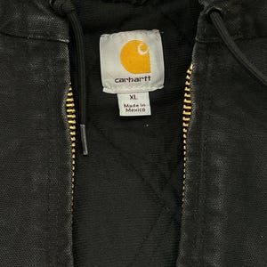 May include: Close-up of a black Carhartt jacket with a gold zipper and a hood. The jacket has a quilted lining and a tag that reads "Carhartt" with the XL size and "Made in Mexico" text.