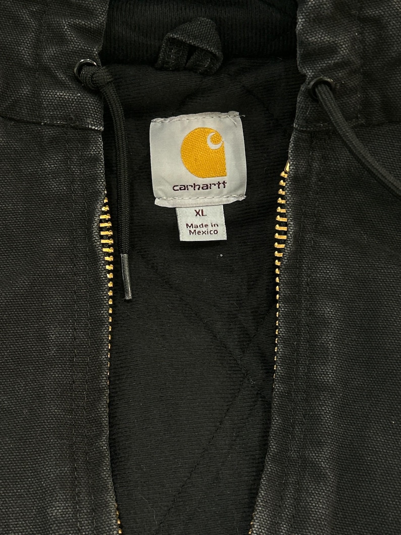 May include: Close-up of a black Carhartt jacket with a gold zipper and a hood. The jacket has a quilted lining and a tag that reads "Carhartt" with the XL size and "Made in Mexico" text.