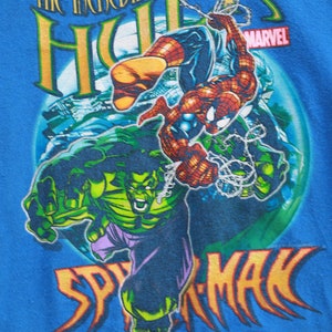 Puede incluir: Una camiseta azul con un estampado gr&aacute;fico de Hulk y Spider-Man. El texto "The Incredible Hulk" y "Spider-Man" est&aacute; impreso en amarillo. El logotipo de Marvel est&aacute; impreso en rojo y blanco.