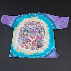 May include: A tie-dye t-shirt featuring a purple bear with a red robe and a crown. The bear is standing in a doorway with a stained glass window above. The shirt is a mix of blue, green, purple, and yellow.