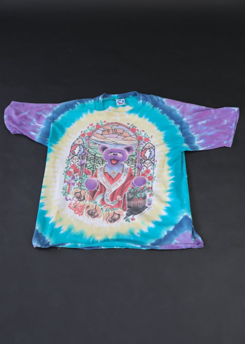 May include: A tie-dye t-shirt featuring a purple bear with a red robe and a crown. The bear is standing in a doorway with a stained glass window above. The shirt is a mix of blue, green, purple, and yellow.