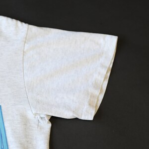 May include: A white t-shirt with a blue and white graphic print. The shirt has a small tear on the sleeve.