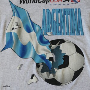 May include: A gray t-shirt with a graphic of a soccer ball breaking apart with a blue and white flag behind it. The text "World Cup USA 94" and "ARGENTINA" are printed on the shirt.