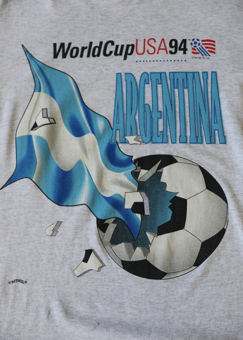 May include: A gray t-shirt with a graphic of a soccer ball breaking apart with a blue and white flag behind it. The text "World Cup USA 94" and "ARGENTINA" are printed on the shirt.