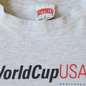 May include: Gray t-shirt with the text "World Cup USA" printed in black and red. The shirt has a tag that reads "Nutmeg" and "Made in U.S.A."