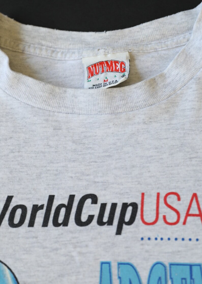 May include: Gray t-shirt with the text "World Cup USA" printed in black and red. The shirt has a tag that reads "Nutmeg" and "Made in U.S.A."