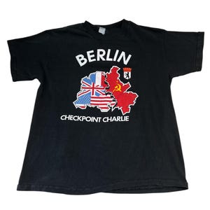 Könnte beinhalten: Schwarzes T-Shirt mit einer weißen Grafik einer Karte von Berlin, Deutschland. Die Karte ist in Abschnitte unterteilt, die das ehemalige Ost- und West-Berlin darstellen, mit den Flaggen von Großbritannien, den Vereinigten Staaten und der Sowjetunion. Der Text "BERLIN" und "CHECKPOINT CHARLIE" ist auf dem Shirt aufgedruckt.