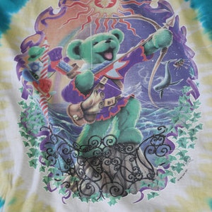 May include: A tie-dye t-shirt featuring a green bear with a bow and arrow, standing on a stone fence with a sun and a dolphin in the background. The shirt has a tie-dye pattern in shades of blue, green, and yellow.