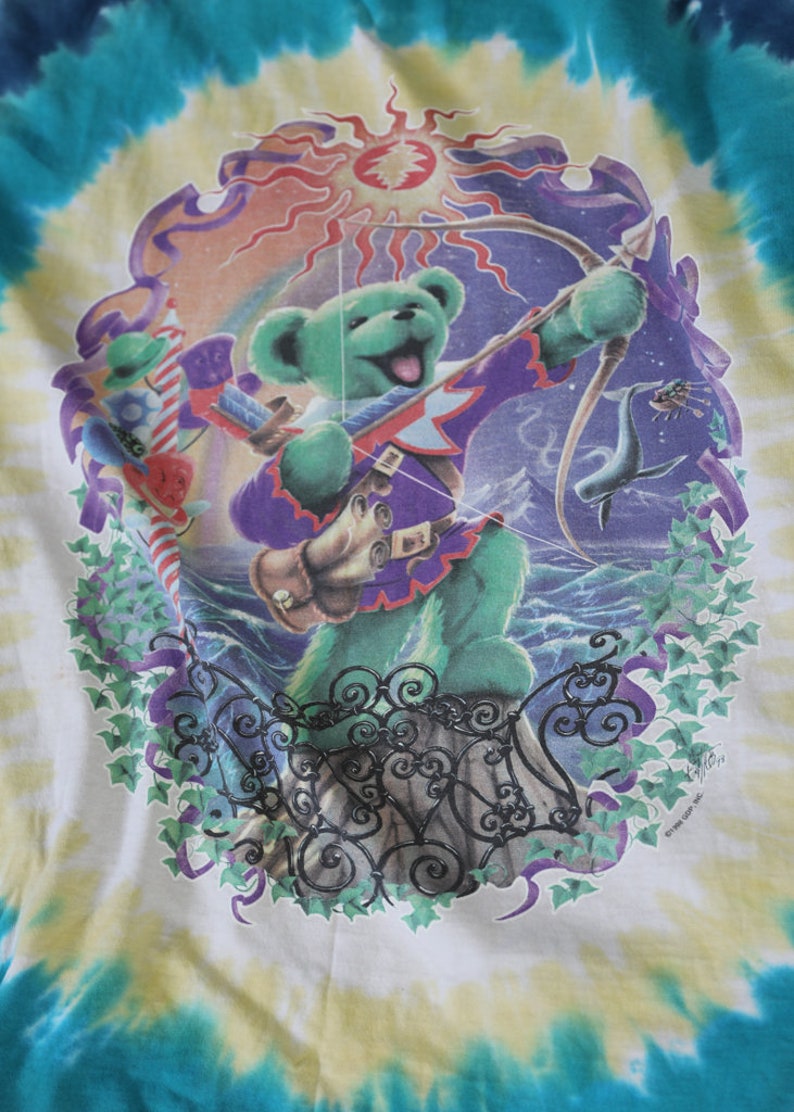 May include: A tie-dye t-shirt featuring a green bear with a bow and arrow, standing on a stone fence with a sun and a dolphin in the background. The shirt has a tie-dye pattern in shades of blue, green, and yellow.