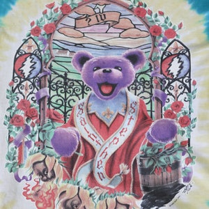 May include: A tie-dye t-shirt featuring a purple bear wearing a red robe with the text "God is in the house" on it. The bear is standing in front of a stained glass window with a rose garden surrounding it. The shirt has a vintage look and feel.