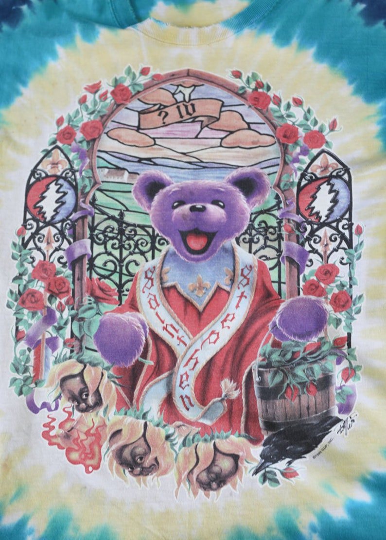 May include: A tie-dye t-shirt featuring a purple bear wearing a red robe with the text "God is in the house" on it. The bear is standing in front of a stained glass window with a rose garden surrounding it. The shirt has a vintage look and feel.