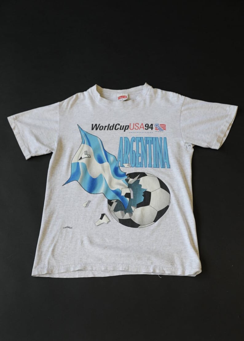 May include: A gray t-shirt with a graphic of a soccer ball being broken apart by the Argentinian flag. The text "WorldCupUSA94" and "ARGENTINA" are printed on the shirt.
