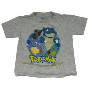 Puede incluir: Una camiseta gris de manga corta con un gráfico colorido de personajes de Pokémon. El diseño incluye la palabra "Pokémon" en amarillo y azul, junto con los nombres "Wartortle", "Blastoise" y "Squirtle".