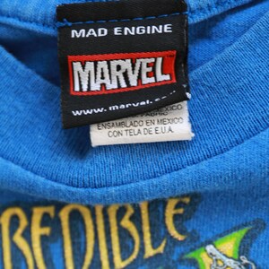 Puede incluir: Una camiseta azul con un gr&aacute;fico amarillo y verde que dice "Incredible Hulk". La camiseta tiene una etiqueta negra con el texto "Mad Engine", "Marvel" y "www.marvel.com".