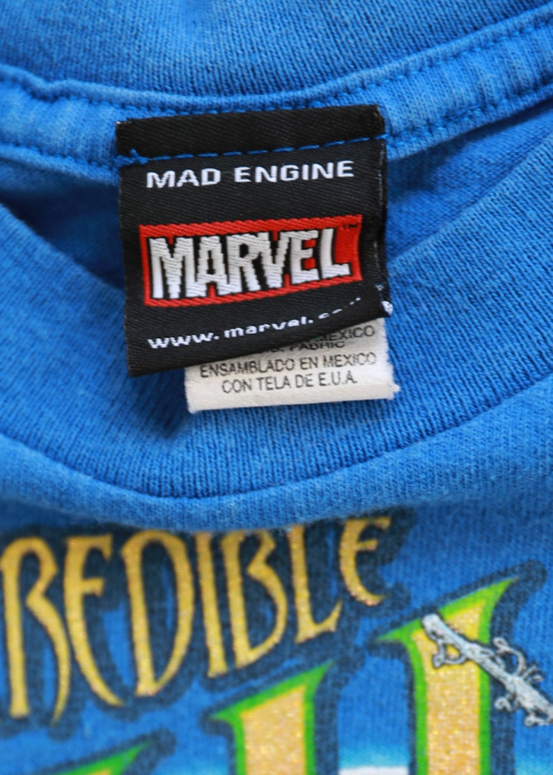 Puede incluir: Una camiseta azul con un gr&aacute;fico amarillo y verde que dice "Incredible Hulk". La camiseta tiene una etiqueta negra con el texto "Mad Engine", "Marvel" y "www.marvel.com".