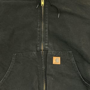 May include: Black hooded jacket with a full-length brass zipper and a front pocket. The jacket features a brown square patch with the Carhartt logo. The jacket has a drawstring hood and is made of a durable, workwear-style fabric.