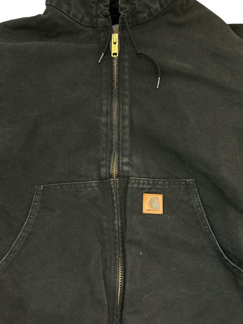 May include: Black hooded jacket with a full-length brass zipper and a front pocket. The jacket features a brown square patch with the Carhartt logo. The jacket has a drawstring hood and is made of a durable, workwear-style fabric.