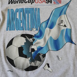 May include: A gray t-shirt with a graphic of a soccer ball being broken apart by a blue and white Argentinian flag. The text "World Cup USA 94" and "ARGENTINA" are printed on the shirt.