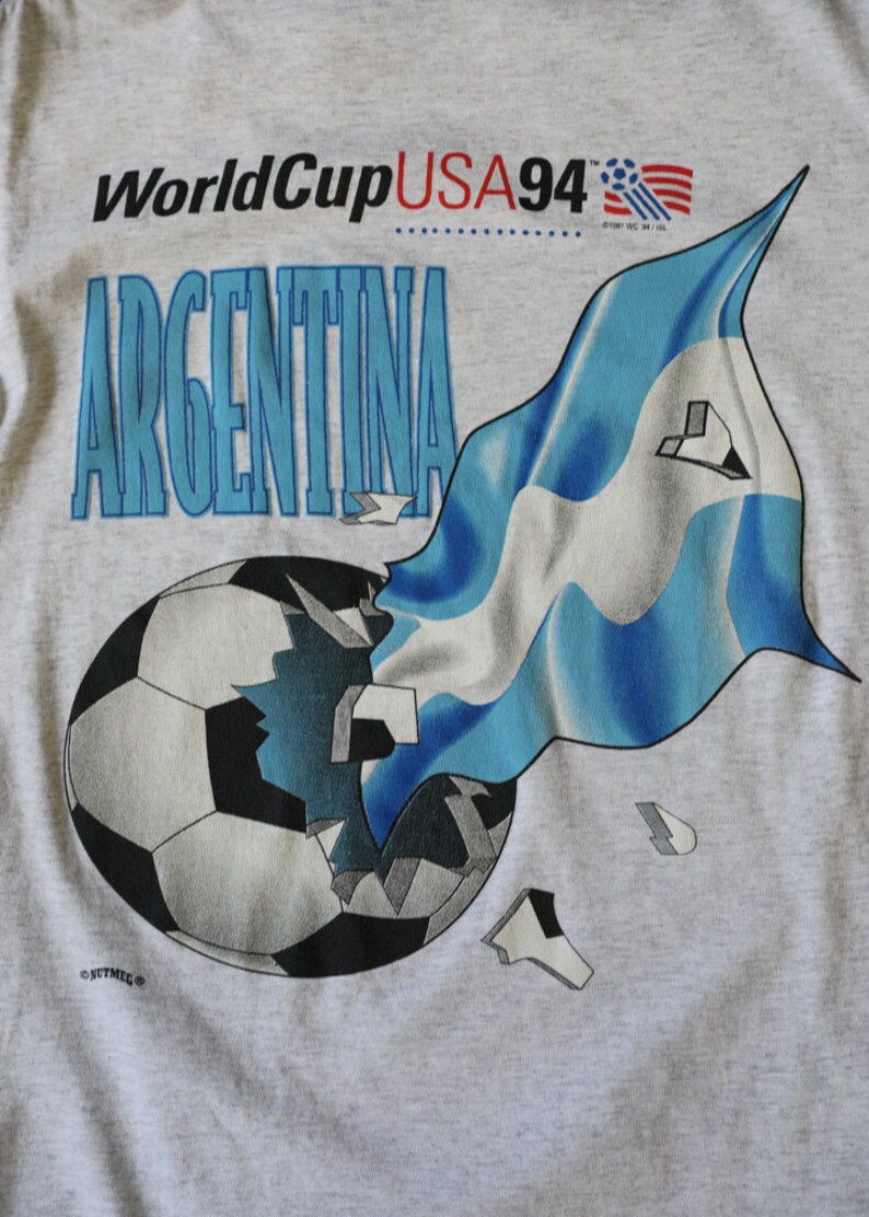 May include: A gray t-shirt with a graphic of a soccer ball being broken apart by a blue and white Argentinian flag. The text "World Cup USA 94" and "ARGENTINA" are printed on the shirt.