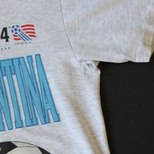 May include: A gray t-shirt with a blue and white graphic of a soccer ball and the word "Argentina". The graphic also includes a small American flag and the text "'94".