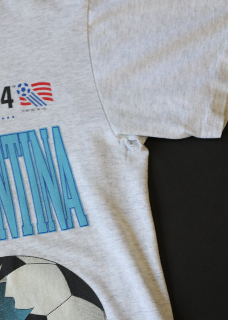 May include: A gray t-shirt with a blue and white graphic of a soccer ball and the word "Argentina". The graphic also includes a small American flag and the text "'94".