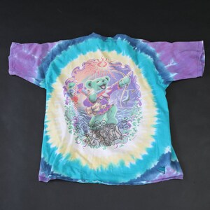 May include: A tie-dye t-shirt with a colorful graphic of a bear playing a guitar. The bear is surrounded by a sun, stars, and other imagery. The shirt is a vibrant blue, purple, and yellow.