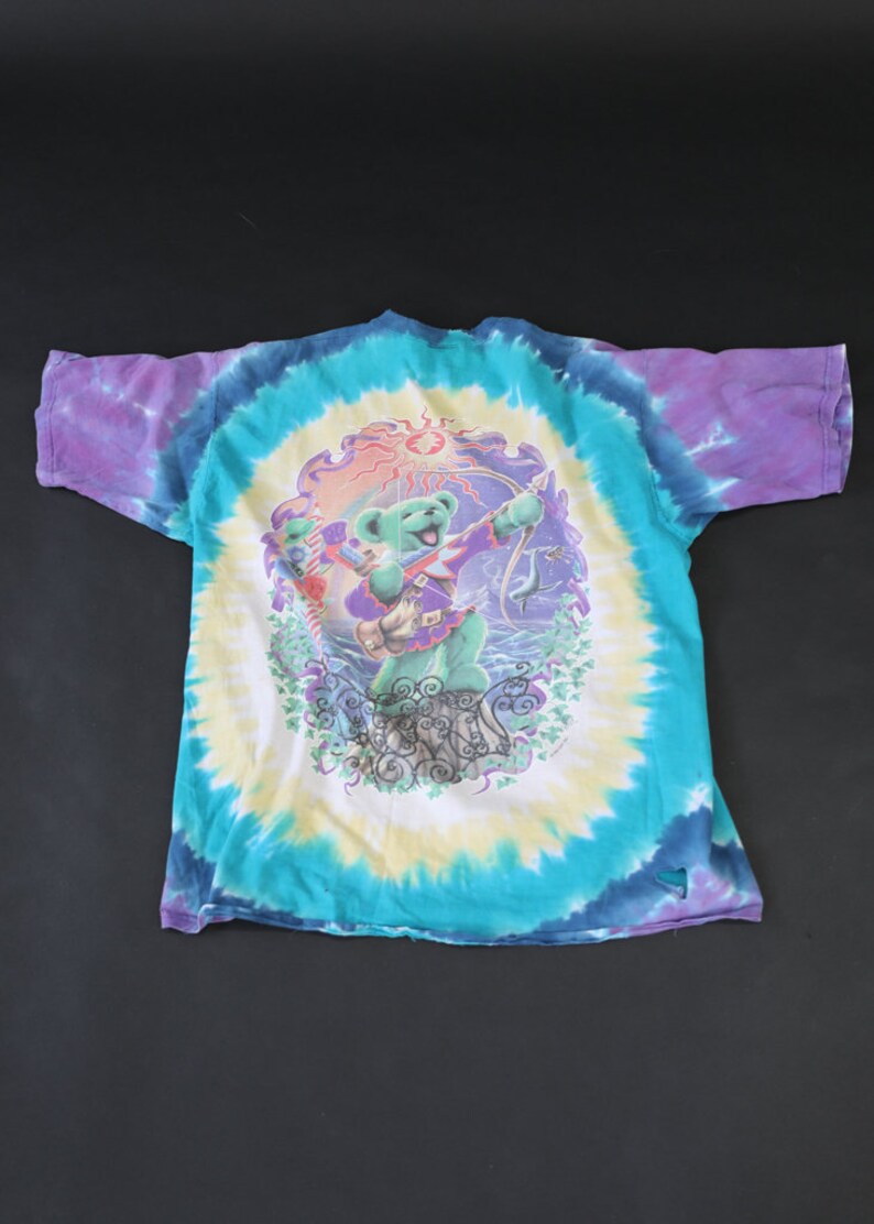 May include: A tie-dye t-shirt with a colorful graphic of a bear playing a guitar. The bear is surrounded by a sun, stars, and other imagery. The shirt is a vibrant blue, purple, and yellow.