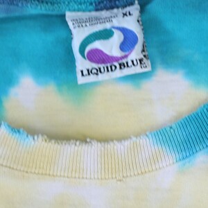 May include: A close-up of a white tag with the text "LIQUID BLUE" in blue letters. The tag is attached to a blue, yellow, and white tie-dye shirt. The shirt is size XL.