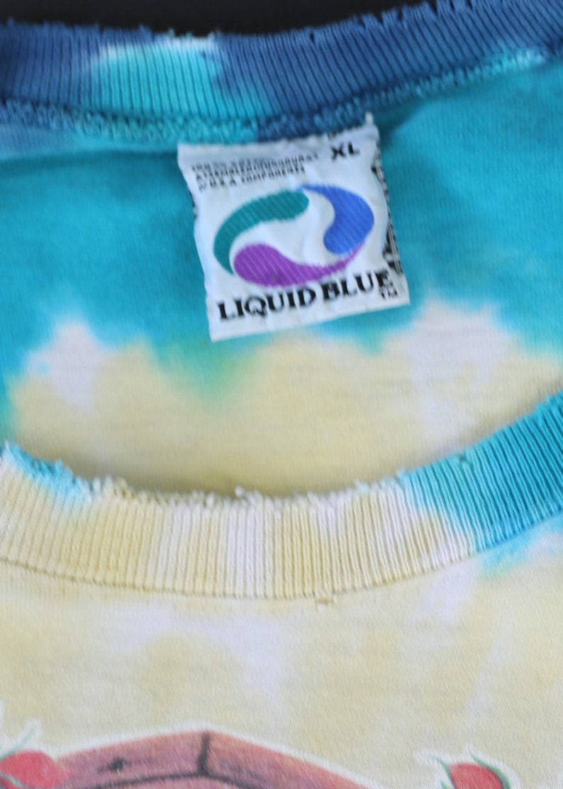 May include: A close-up of a white tag with the text "LIQUID BLUE" in blue letters. The tag is attached to a blue, yellow, and white tie-dye shirt. The shirt is size XL.