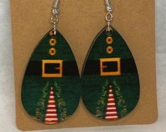 Puede incluir: Un par de pendientes colgantes de temática elfo verde y dorado. Los pendientes están hechos de madera y tienen forma de lágrima. El elfo lleva un gorro verde, un cinturón negro con hebilla dorada y una camisa a rayas rojas y blancas.