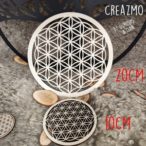 K&ouml;nnte beinhalten: Zwei Holzuntersetzer mit einem geometrischen Blumen des Lebens-Design. Der gr&ouml;&szlig;ere Untersetzer hat einen Durchmesser von 20 cm, der kleinere 10 cm. Die Untersetzer sind hell und liegen auf einer strukturierten, grauen Oberfl&auml;che.