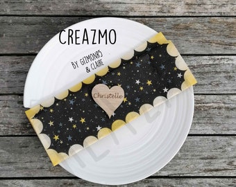 Könnte beinhalten: Ein weißer Teller mit einer schwarz-gelb gemusterten Serviette mit Sternen und einem Holzherz mit dem Namen "Christelle". Die Serviette hat einen gewellten Rand und den Text "CREAZMO BY GIZMONR5 & CLAIRE".