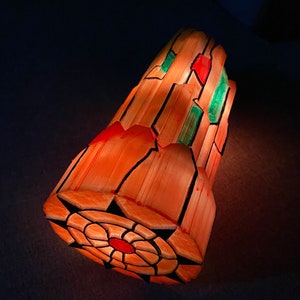 Puede incluir: Una pieza de arte artesanal e iluminada con un diseño geométrico. El objeto es principalmente naranja con acentos rojos y verdes, y contornos negros. La base presenta un patrón circular con un elemento central rojo. La pieza está iluminada desde dentro, creando un brillo cálido.