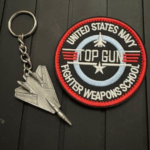 Könnte beinhalten: Ein silberfarbener Metall-Schlüsselanhänger in Form eines Kampfflugzeugs, neben einem runden Aufnäher. Der Aufnäher zeigt den Text "UNITED STATES NAVY TOP GUN FIGHTER WEAPONS SCHOOL" in Weiß und Rot auf schwarzem Hintergrund.