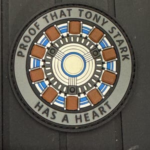 May include: A round, gray patch with a brown and blue Iron Man-style arc reactor design. The text "PROOF THAT TONY STARK HAS A HEART" is written around the edge of the patch.