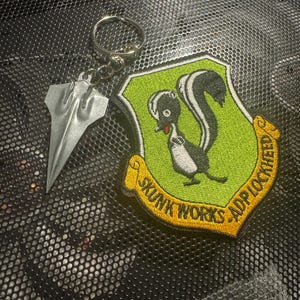 May include: A keychain featuring a silver metal airplane-shaped charm and a patch with a skunk illustration. The patch is green with yellow trim and text that reads "SKUNK WORKS ADP LOCKHEED".