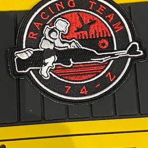 May include: Embroidered patch with the words "RACING TEAM" in a circular design. The patch features a red and black color scheme with a stylized image of a rifle and the number "74". The patch is on a black keyboard.