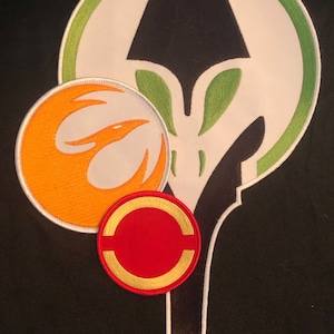 以下が含まれることがあります： さまざまな形と色の刺繍パッチ。黒いデザインの大きな白と緑のパッチ、オレンジと白の円形パッチ、赤と金の円形パッチが含まれています。パッチは黒い背景にあります。