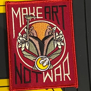 Könnte beinhalten: Roter gestickter Patch mit weißem Umriss. Der Patch zeigt ein stilisiertes Bild eines Mandalorianer-Helms mit Blumen und dem Text "MAKE ART NOT WAR".
