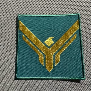 May include: A green fabric patch with a gold embroidered eagle design. The eagle is facing forward with its wings spread out.