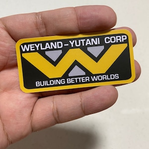 Peut inclure: Un autocollant noir et jaune avec le texte "Weyland-Yutani Corp" et "Building Better Worlds". L'autocollant présente un logo "W" stylisé avec trois triangles gris.