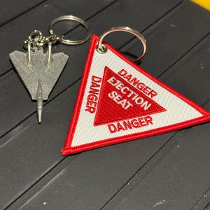 Puede incluir: Un juego de llaveros con un dije de avión de metal plateado y un parche triangular blanco y rojo. El parche dice "DANGER EJECTION SEAT" en letras rojas.