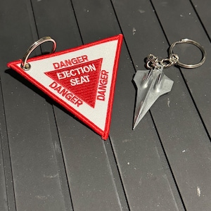 May include: Two keychains. One is a red, white, and black embroidered patch in a triangle shape with the words "DANGER EJECTION SEAT" and "DANGER". The other is a silver-colored metal airplane-shaped keychain.