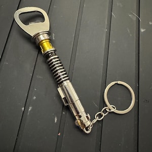 Può includere: Un apribottiglie portachiavi in ​​argento a forma di spada laser. Il portachiavi ha un anello dorato attorno all'apribottiglie.