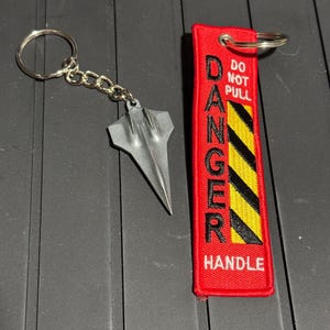 May include: A keychain with a silver metal airplane charm and a red tag. The tag has the words "DANGER" and "HANDLE" in black, with yellow and black diagonal stripes. The text "DO NOT PULL" is also on the tag.