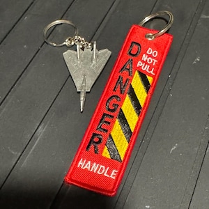 Könnte beinhalten: Ein roter, bestickter Schlüsselanhänger mit dem Wort "DANGER" in Schwarz und gelben Diagonalstreifen. Das Etikett sagt auch "DO NOT PULL" und "HANDLE". Ein silberfarbener Metall-Schlüsselanhänger in Flugzeugform ist befestigt.